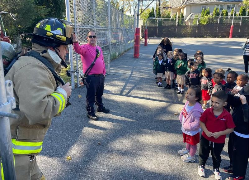 A firefighter talks to a group of young children, with an adult in a pink shirt observing. They're likely learning about fire safety.