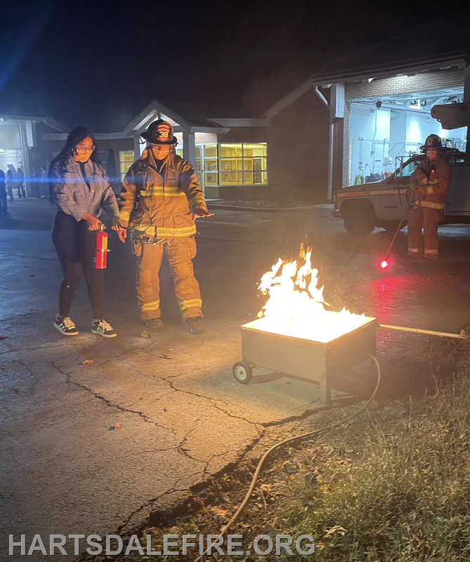 A firefighter instructs a person with a fire extinguisher near a controlled fire at night. Another firefighter is in the background.