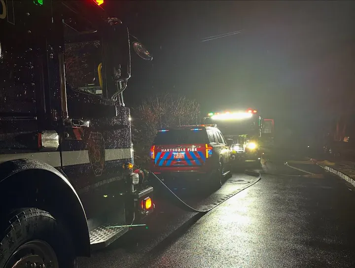 A fire truck and emergency vehicle are parked at night, likely responding to an incident. Rain is visible on the surfaces.