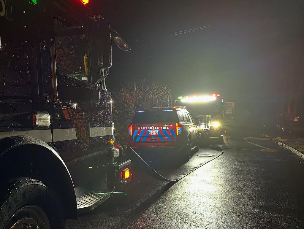 A fire truck and emergency vehicle are parked at night, likely responding to an incident. Rain is visible on the surfaces.