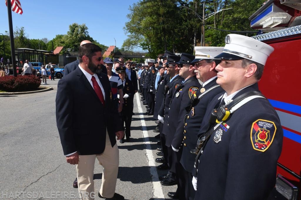 A line of firefighters in uniform stands in formation, facing a man in a suit, near fire trucks on a sunny day.