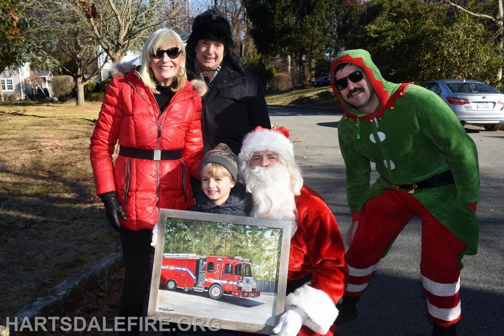 A festive group poses outdoors, featuring Santa and an elf, with a child holding a painting of a fire truck.