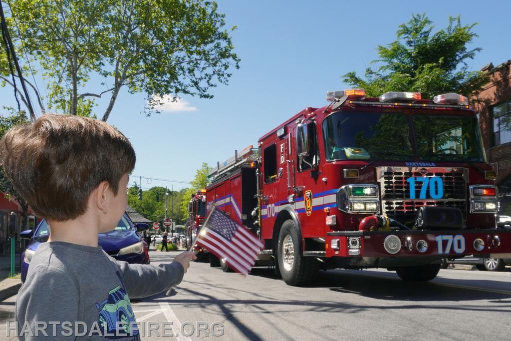 A child waves an American flag at a red fire truck driving by on a sunny street.