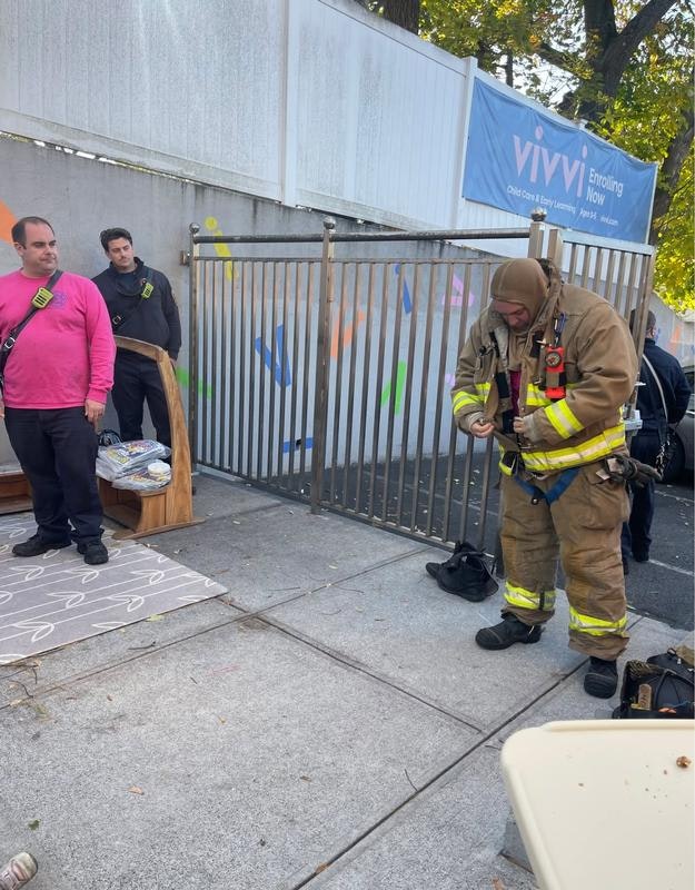 A firefighter prepares equipment while two other individuals look on near a gated area with colorful graffiti.