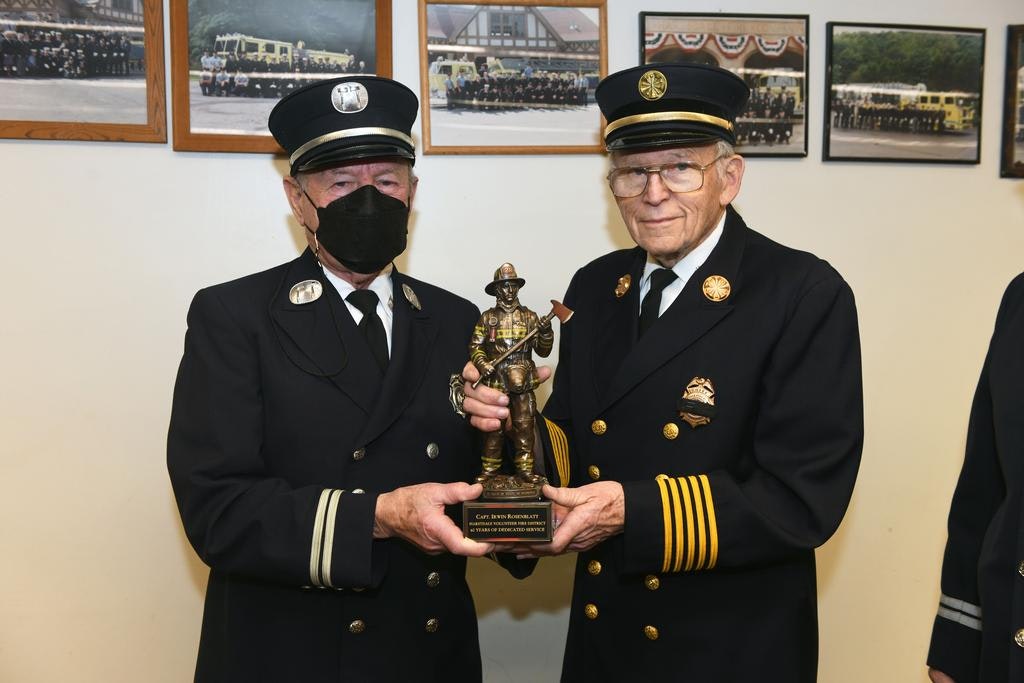 Two uniformed individuals holding a firefighter statue, standing in front of framed photographs.
