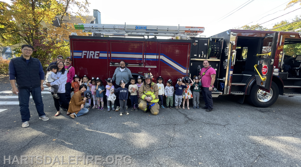 A group of children and adults pose by a fire truck, with some kids wearing firefighter helmets, during an outdoor event.