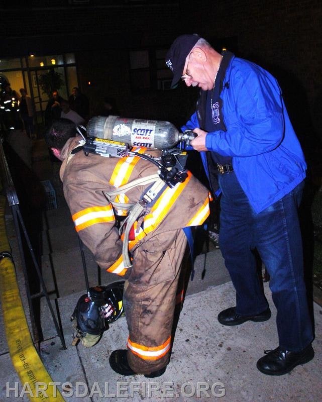 A firefighter in gear is receiving assistance from another person, possibly adjusting equipment, in a nighttime setting.