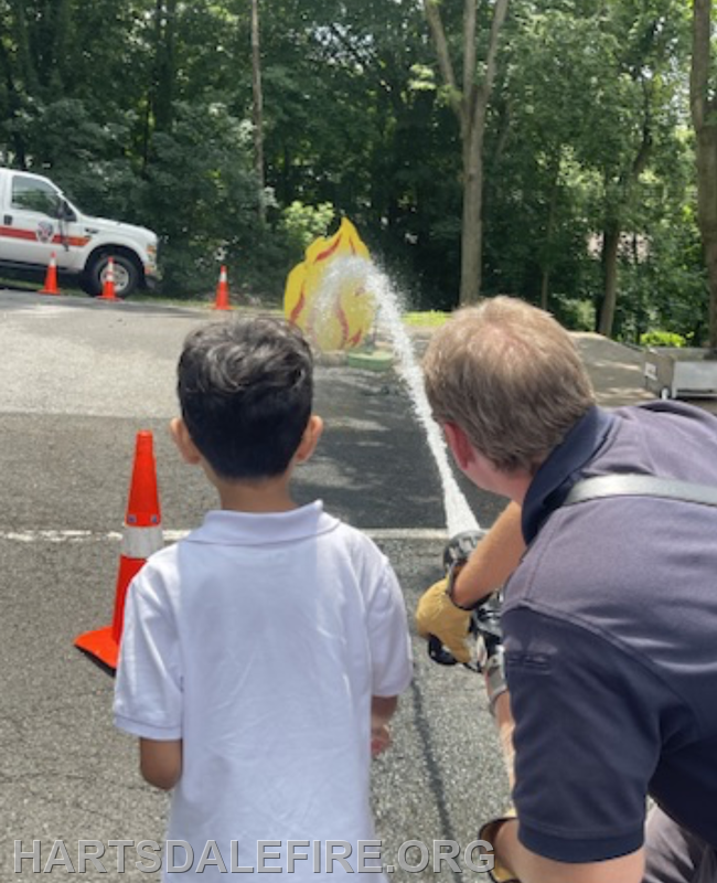 A child watches as a firefighter sprays water at a flame-shaped target during a safety demonstration.