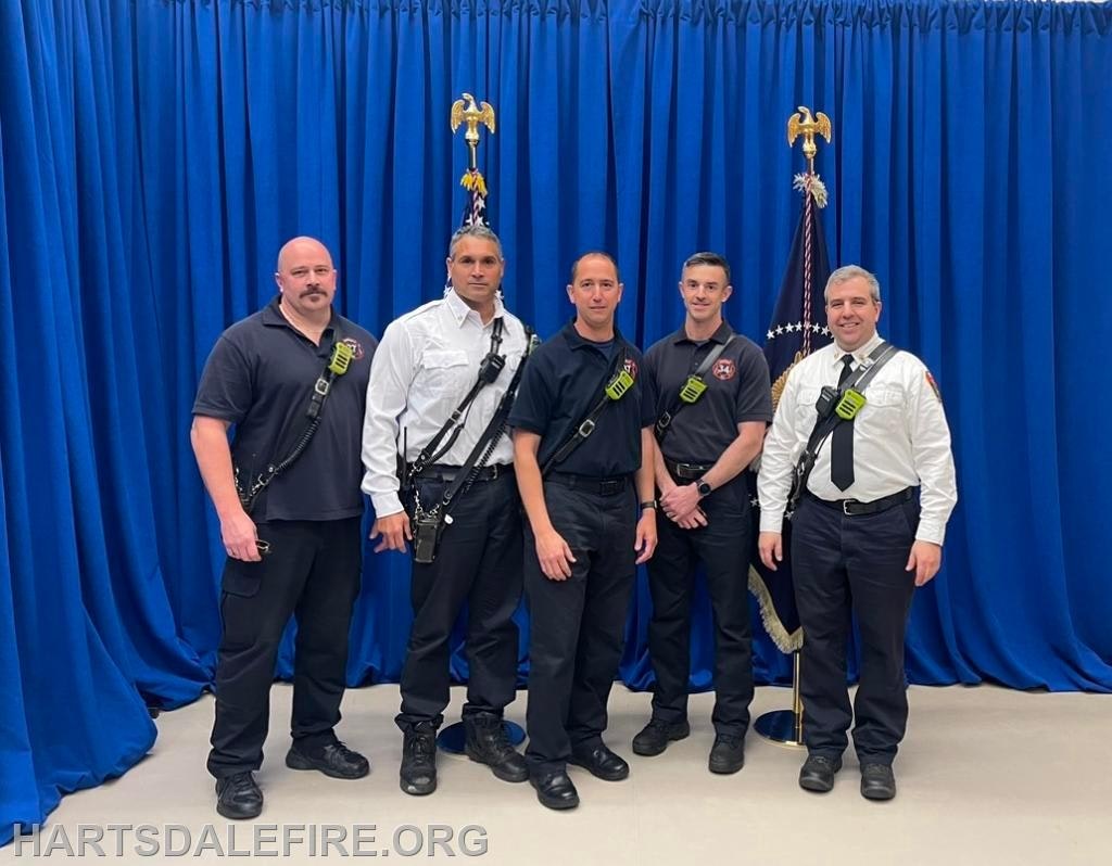 A group of five men, likely firefighters, standing in front of blue curtains with flags behind them.