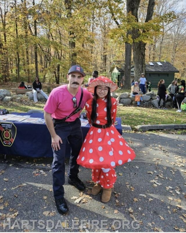 A man in a pink shirt poses with a girl in a polka-dot costume during a community event in a park.