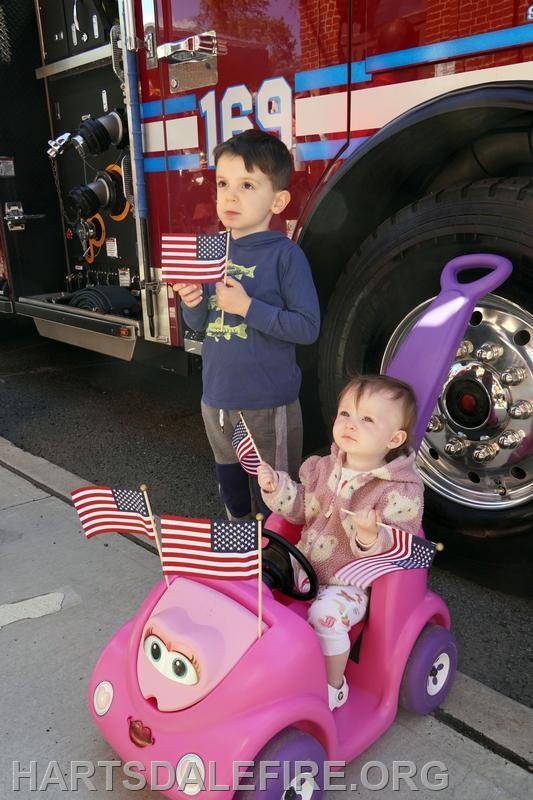 Two young children hold American flags in front of a fire truck, with one sitting in a pink toy car.