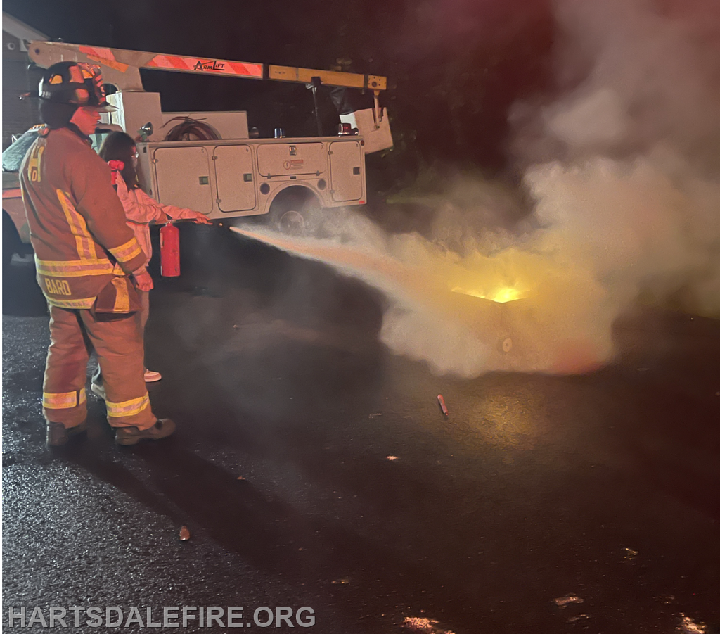 A firefighter and a person are using a fire extinguisher to put out a small fire at night, with smoke billowing around them.