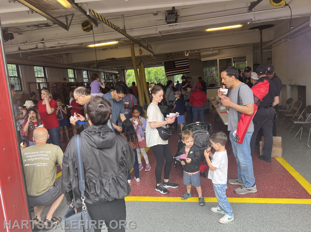 A community event at a fire station with families gathering, enjoying snacks, and chatting, featuring children and adults.