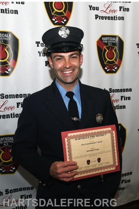 A firefighter in uniform smiles while holding a certificate, posing in front of a backdrop promoting fire prevention.