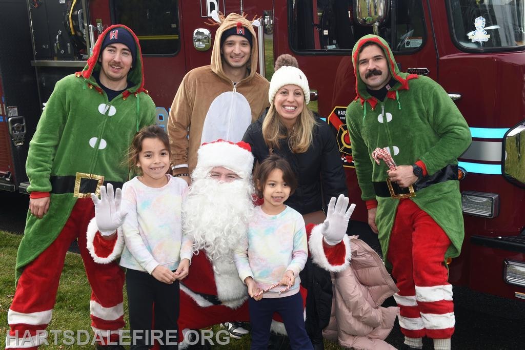 A festive group photo featuring Santa, kids, and individuals in elf costumes by a fire truck, celebrating the holiday spirit.