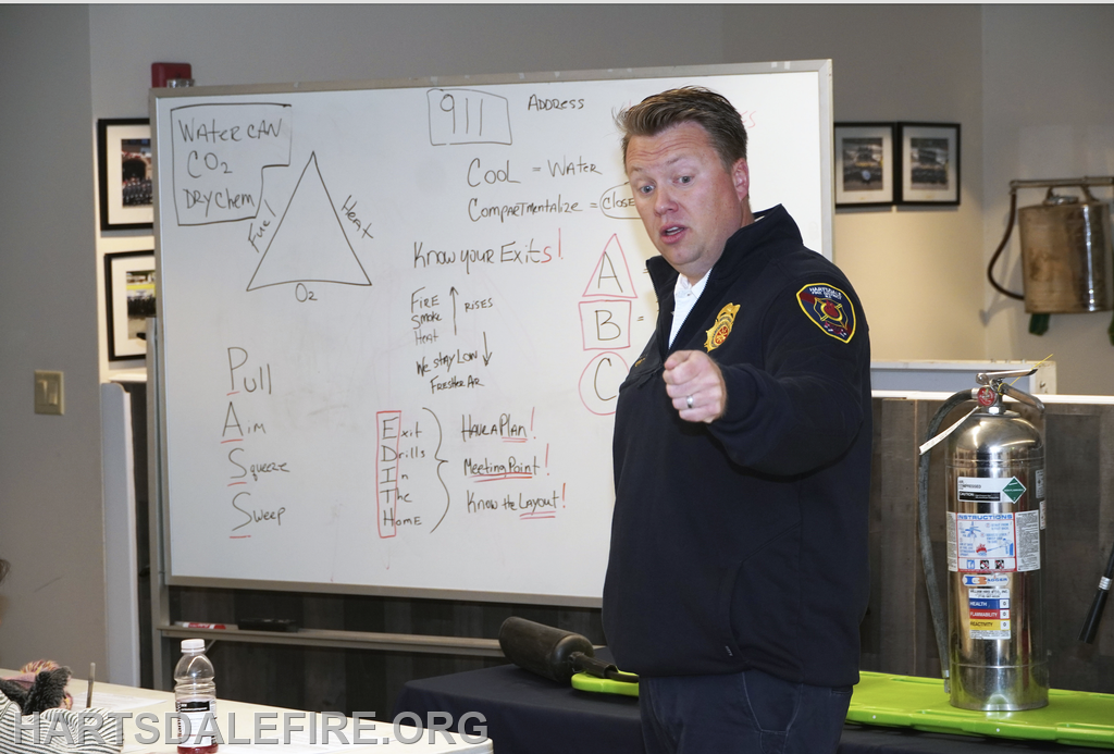 A fire safety presentation with a speaker, whiteboard notes on fire response, and a fire extinguisher in the background.