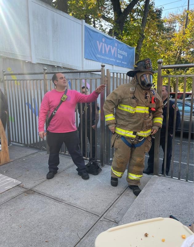 A firefighter in gear stands by a gate while a person in a pink shirt interacts, with others in the background.