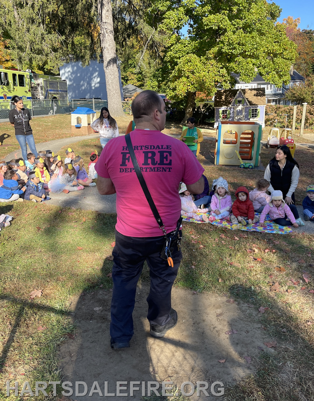 A firefighter in a pink shirt addresses a group of children sitting on the ground in a park, surrounded by adults and play structures.