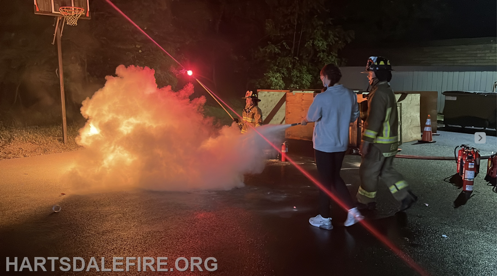 A fire training exercise at night, featuring a person using a fire extinguisher with a firefighter guiding them, amidst smoke.