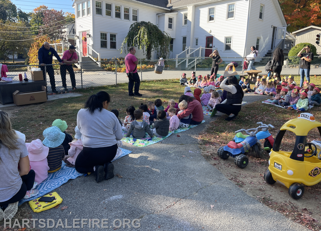 A group of children sit on blankets outside, while adults in varied attire engage with them in a playground setting.