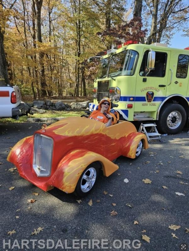 A child in an orange costume sits in a small, flame-painted car near a fire truck, surrounded by autumn trees.