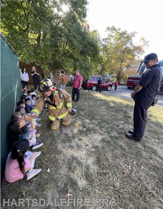 A firefighter interacts with children sitting on the ground, while other adults and emergency vehicles are present nearby.