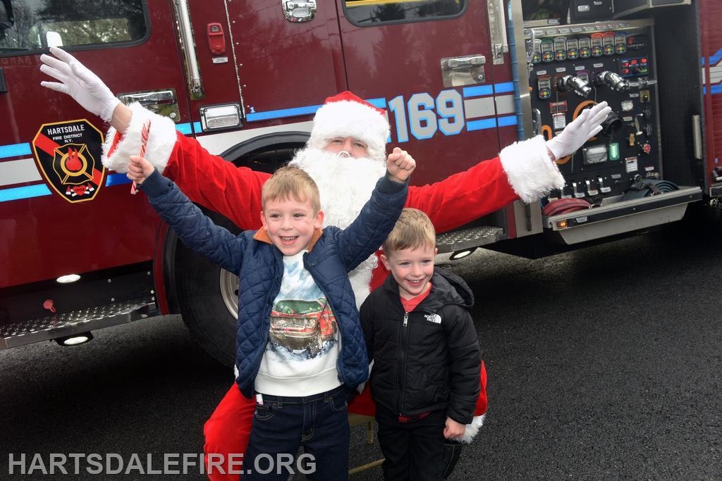 Two kids pose excitedly in front of a fire truck with Santa, celebrating the holiday spirit.