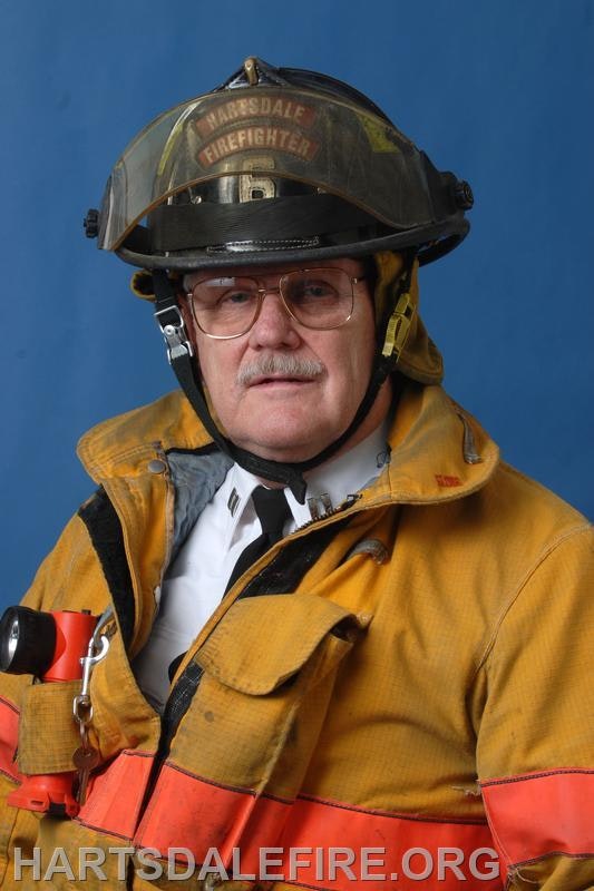 A firefighter in uniform and helmet poses for a portrait against a blue background.