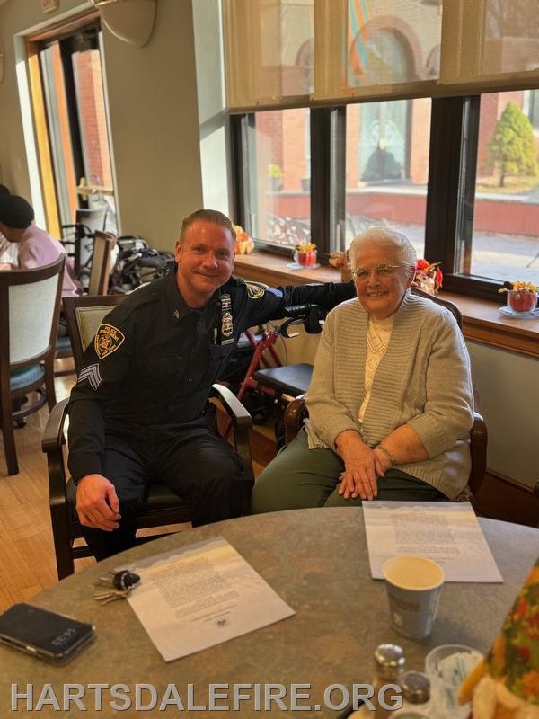 A police officer and an elderly woman smile together at a table, creating a warm, friendly atmosphere in a communal space.