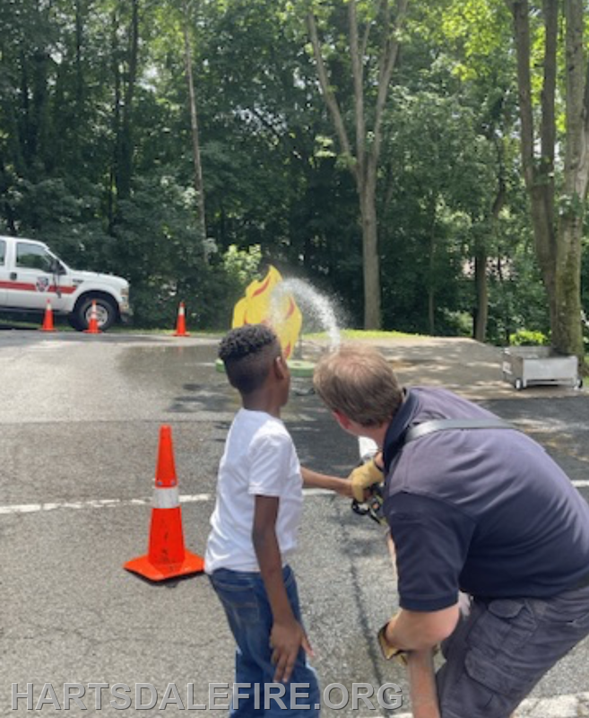 A child is learning to use a fire hose with guidance from a firefighter, aiming at a flame target surrounded by safety cones.