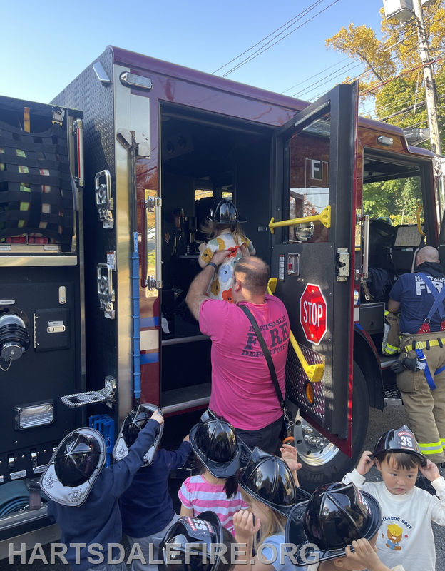 A firefighter helps a child in a costume from a fire truck while other kids in helmets watch excitedly.
