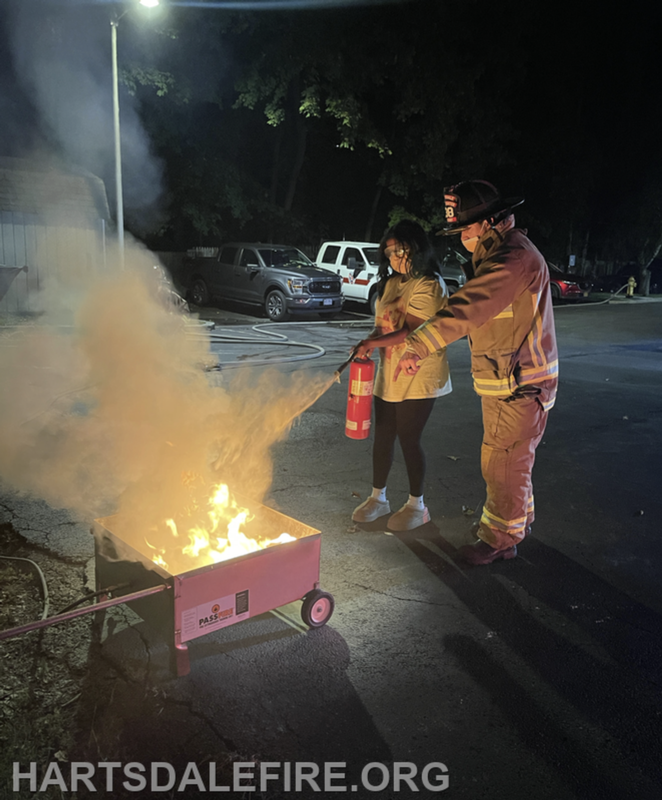 A person in firefighter gear demonstrates using a fire extinguisher on a small fire while someone observes.