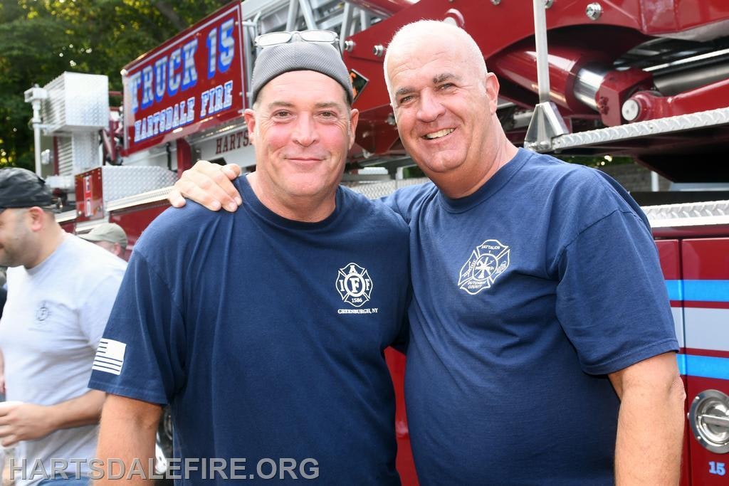 Two smiling firefighters pose together in front of a fire truck, showcasing camaraderie and team spirit.