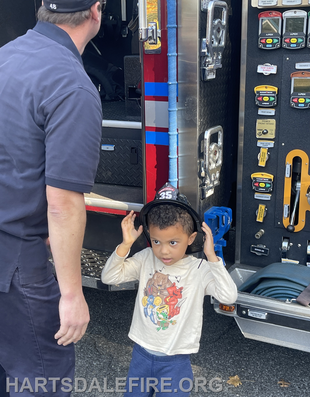 A child wearing a firefighter hat interacts with an adult by a fire truck, exploring the gear and environment.