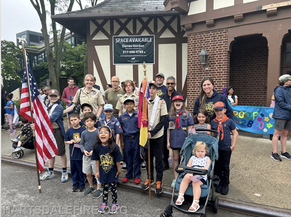 A group photo of children and adults in a parade, many wearing uniforms, holding flags, with a "Space Available" sign nearby.