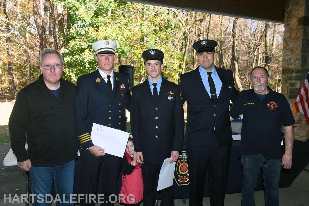 A group of uniformed firefighters and one civilian pose together, with a child peeking out from behind.