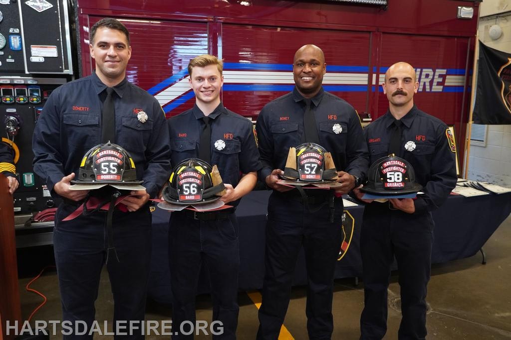Four firefighters in uniform holding helmets, standing in front of a fire truck.
