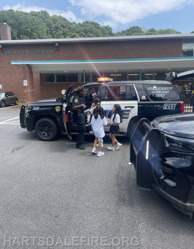 Children interact with a police officer near a school, with a police vehicle in the foreground under a clear sky.
