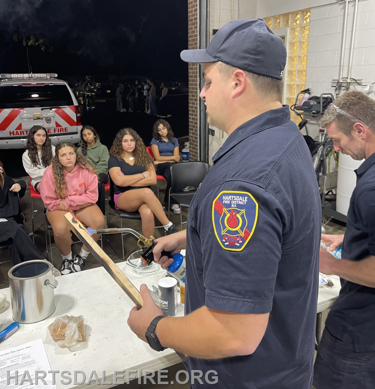 A firefighter demonstrates fire safety techniques to a group of seated young people at a fire district event.