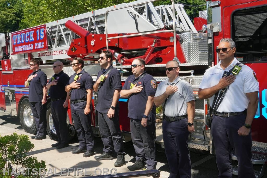 Firefighters standing in a line with hands on their hearts in front of a fire truck, likely paying tribute or honoring an event.