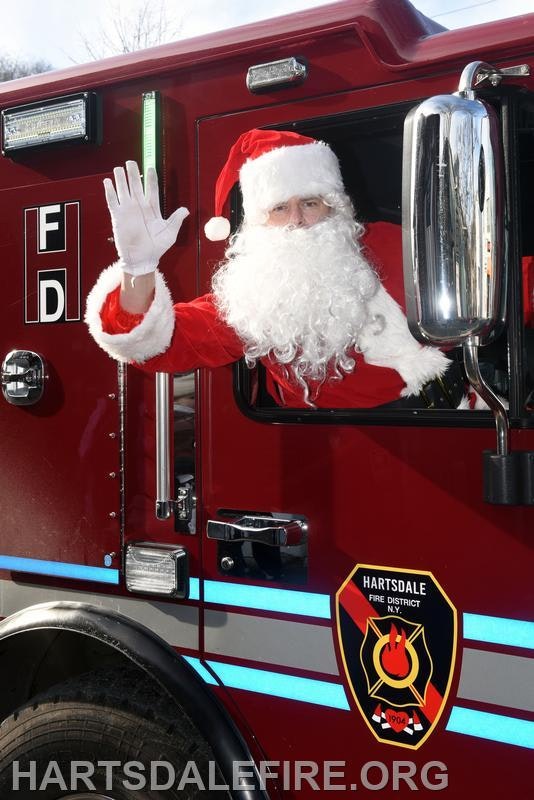A festive Santa Claus is waving from the window of a fire truck, promoting holiday cheer.