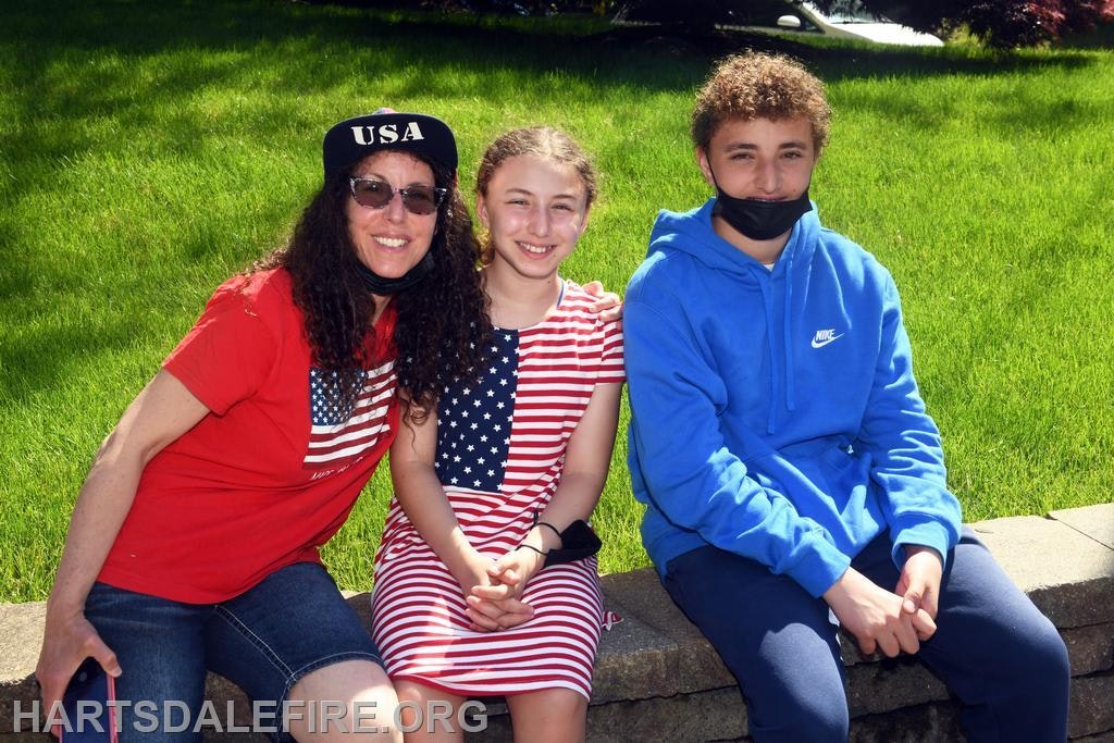Three people sit outside: one in a red USA shirt, one in a striped dress, and one in a blue hoodie.