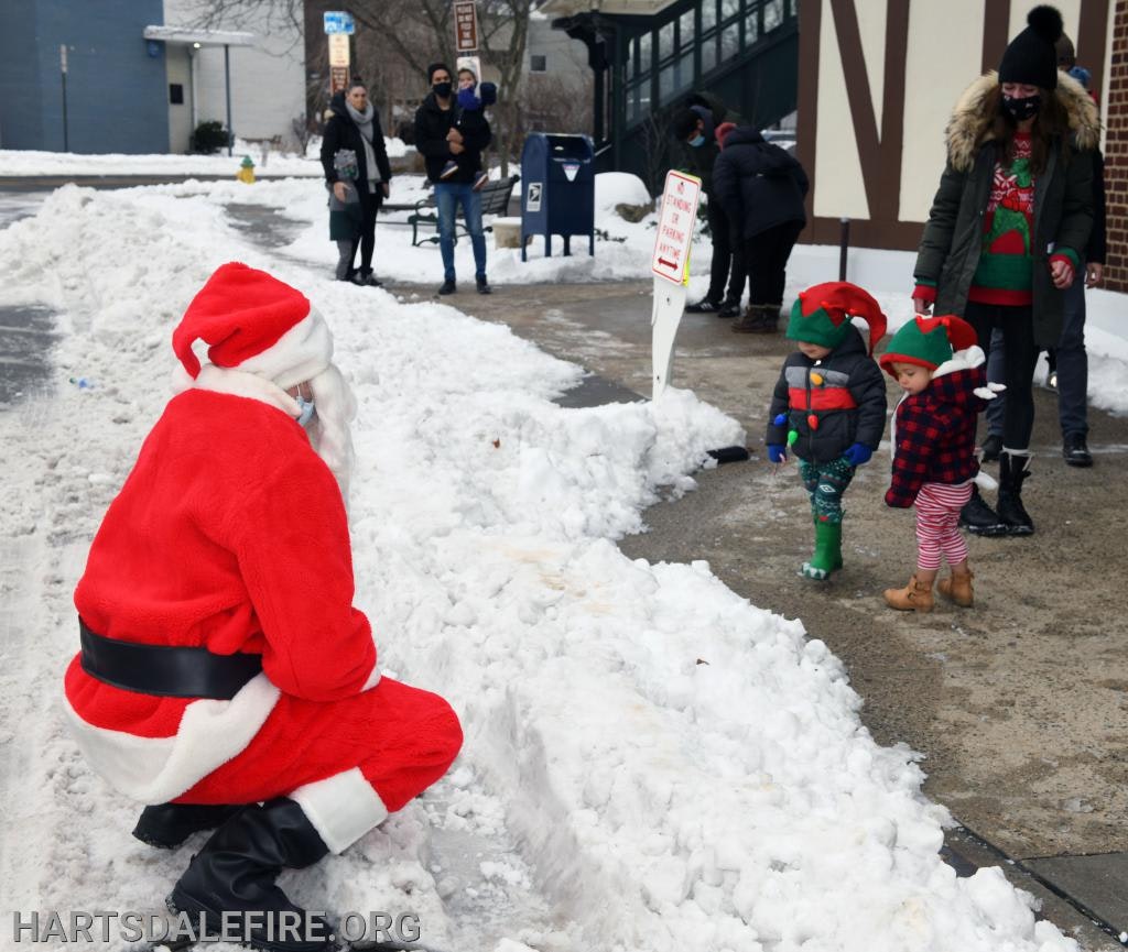 Santa kneeling in snow to greet two children dressed as elves; adults in winter attire are nearby.