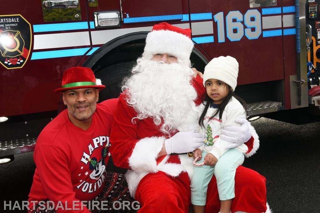A man in a festive outfit and Santa pose with a young girl in winter attire in front of a fire truck. Holiday spirit!