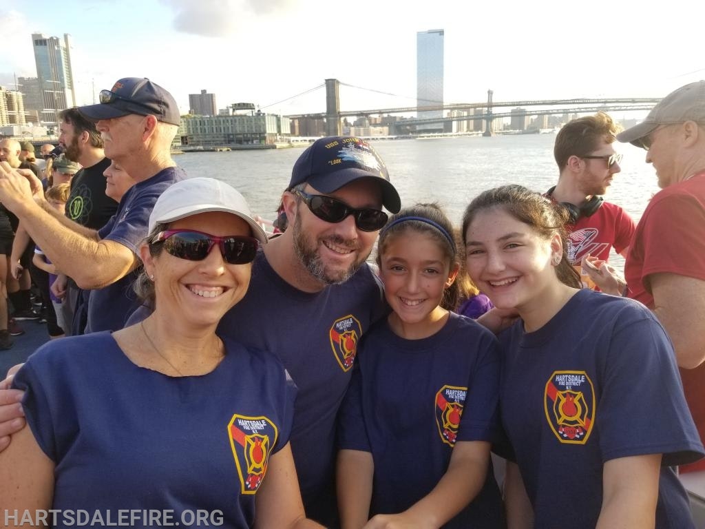 A group of people smiling near a waterfront, some in matching blue shirts with a fire department logo. City skyline and bridge in background.