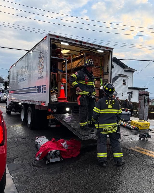 Firefighters are unloading equipment from a fire truck at an incident scene, with a cloudy sky overhead.