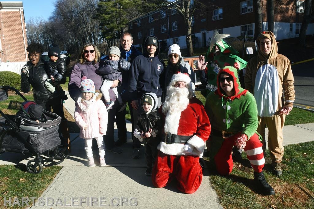 A festive group photo with children, adults, Santa, and holiday-themed costumes outside a building.