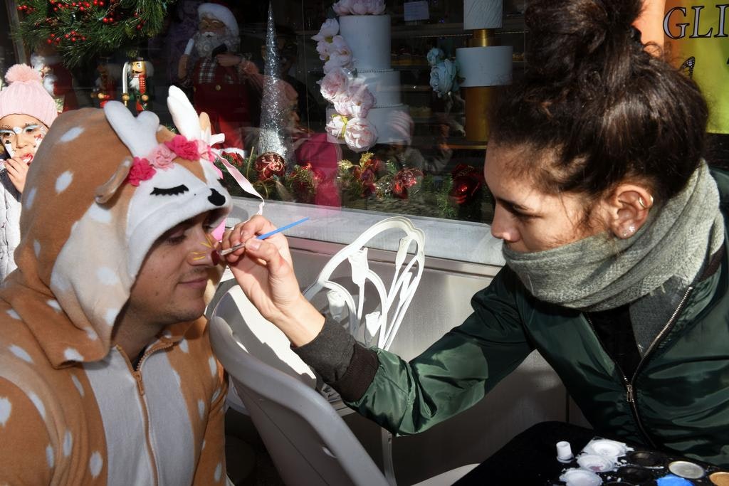 A person in a deer onesie is having their face painted by another person outdoors, surrounded by festive decorations.