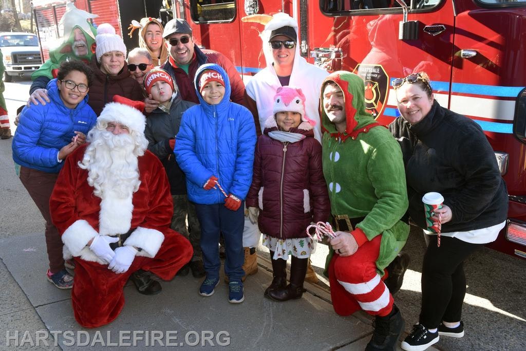 A festive group poses with Santa and an elf near a fire truck, celebrating the holiday season with smiles and candy canes.
