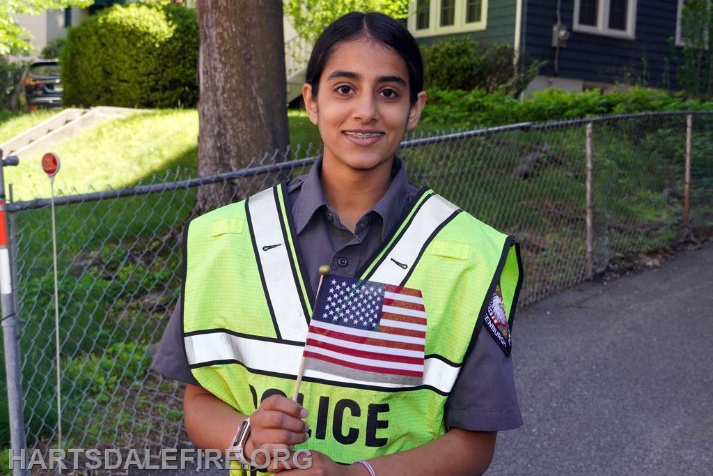 A young individual in a police vest stands outdoors, holding a small American flag, with a tree and houses in the background.